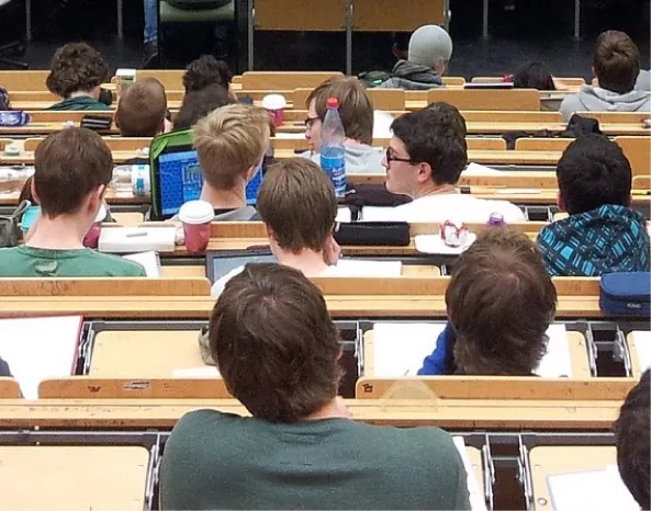 Picture is in the center. Many rows of students are in a classroom. One student has an open laptop on his desk.